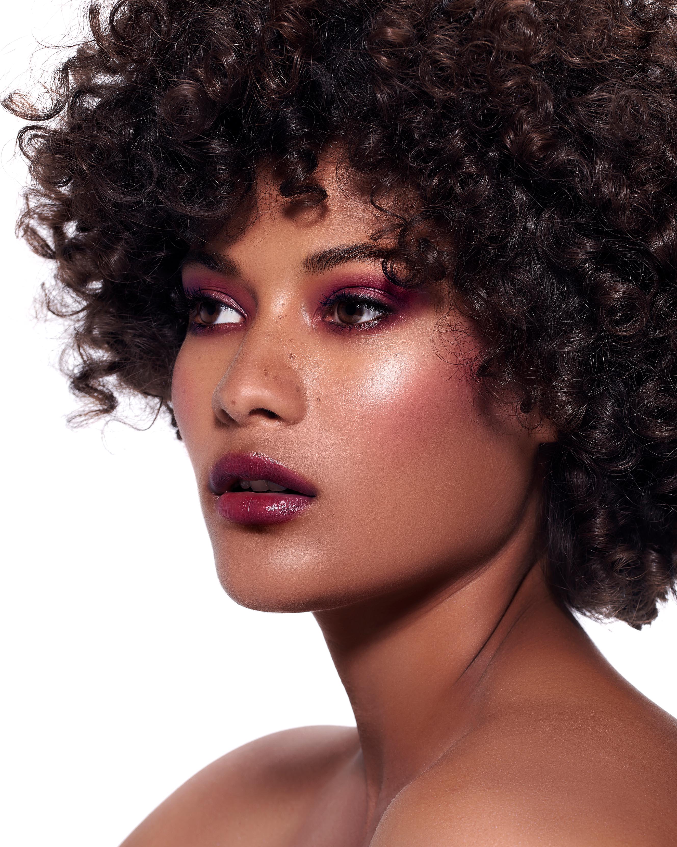 Close-up of a model with medium-deep skin wearing purple makeup on lips, eyes, and cheeks